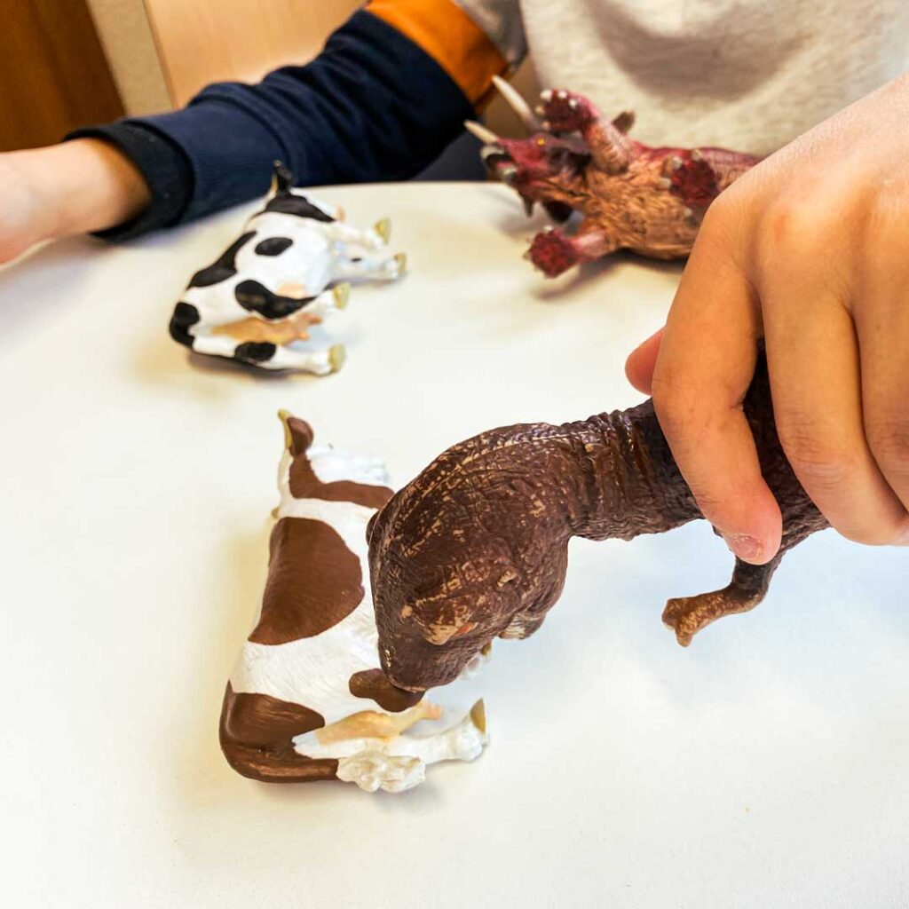 Ein Kind spielt mit Tierfiguren – einem Dino und mehreren Kühen.