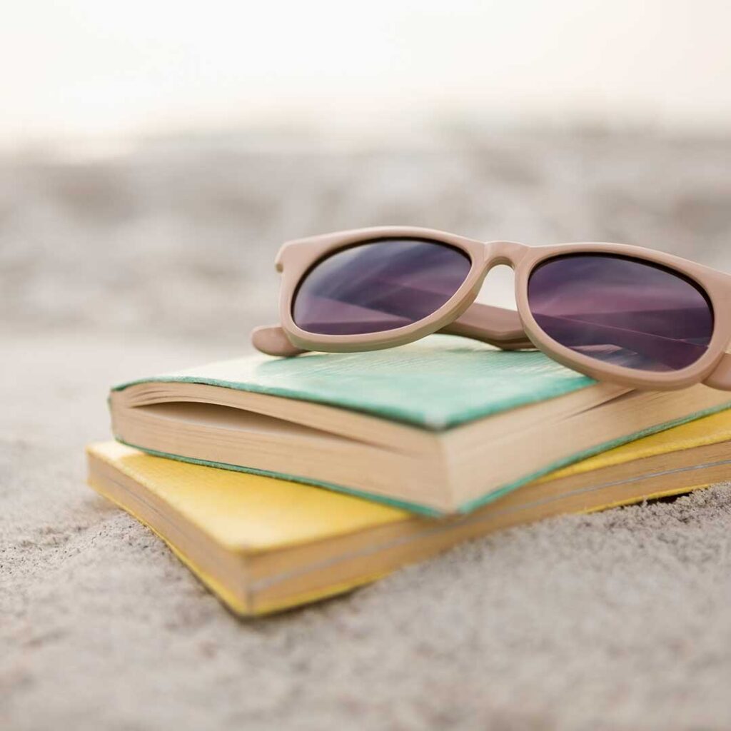 Symbolbild für eine Sommerpause: zwei Bücher liegen im Sandstrand, darauf eine Sonnenbrille